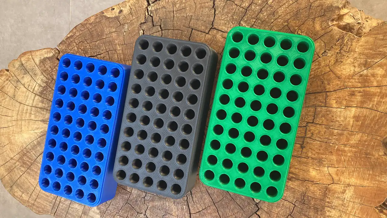 Três suportes retangulares perfurados, um azul, um cinza e um verde, lado a lado sobre um tronco cortado